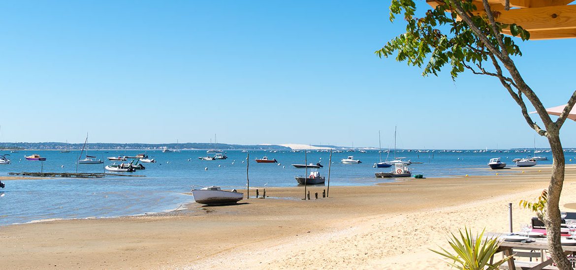 Plage de sable à Arcachon avec des bateaux amarrés et quelques voiliers au loin sur une mer calme, vue depuis une terrasse en bois avec des arbres et des tables de restaurant. Le ciel est bleu sans nuages.