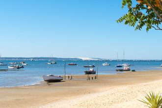 Plage de sable à Arcachon avec des bateaux amarrés et quelques voiliers au loin sur une mer calme, vue depuis une terrasse en bois avec des arbres et des tables de restaurant. Le ciel est bleu sans nuages.