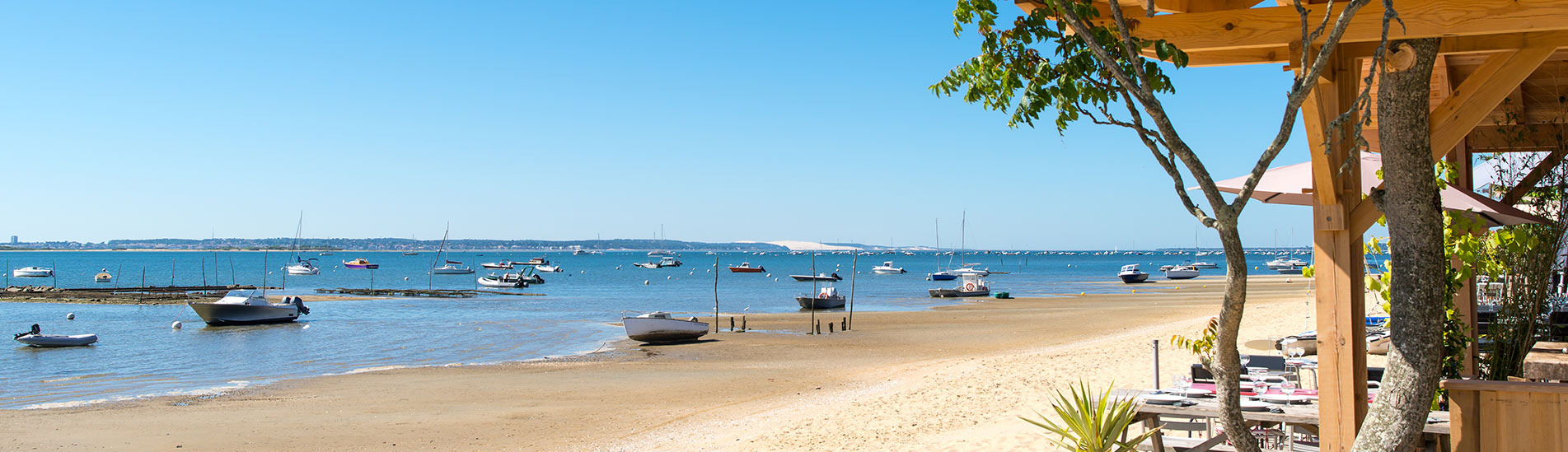 Plage de sable à Arcachon avec des bateaux amarrés et quelques voiliers au loin sur une mer calme, vue depuis une terrasse en bois avec des arbres et des tables de restaurant. Le ciel est bleu sans nuages.