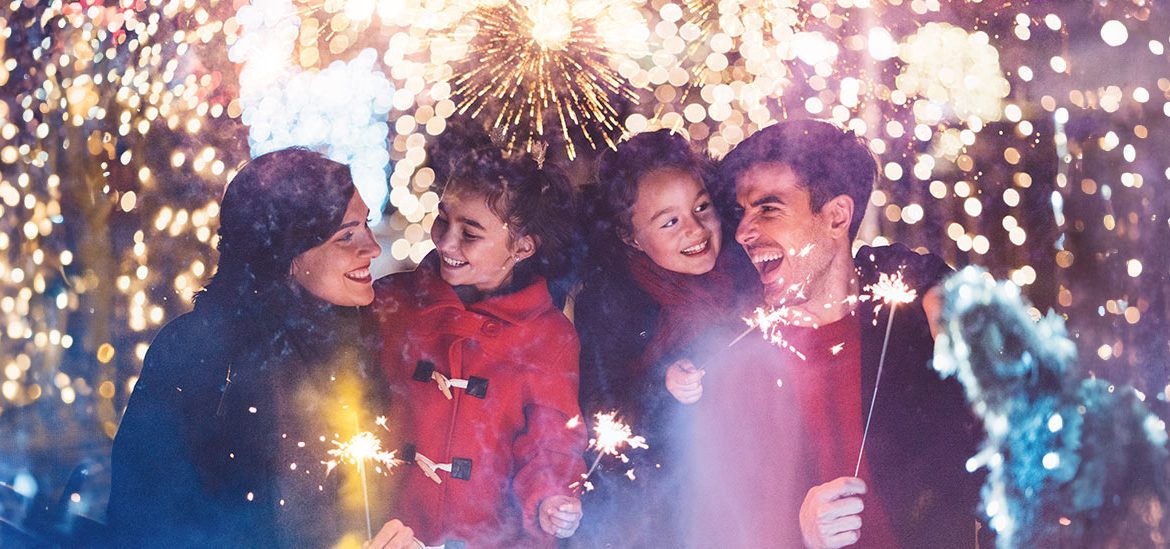 Famille souriante tenant des cierges magiques devant des feux d'artifice éclatants et des lumières festives en arrière-plan. Les parents et leurs deux enfants semblent joyeux et émerveillés.