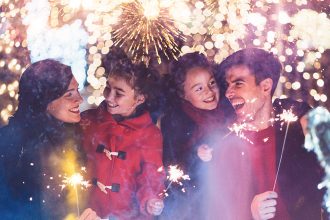 Famille souriante tenant des cierges magiques devant des feux d'artifice éclatants et des lumières festives en arrière-plan. Les parents et leurs deux enfants semblent joyeux et émerveillés.