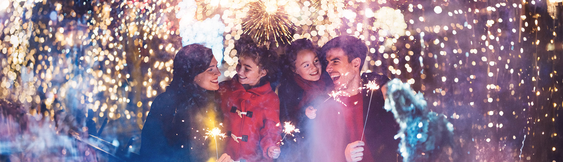 Famille souriante tenant des cierges magiques devant des feux d'artifice éclatants et des lumières festives en arrière-plan. Les parents et leurs deux enfants semblent joyeux et émerveillés.