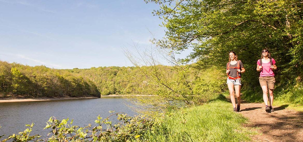 Deux randonneurs marchant sur un sentier en bord de rivière, entourés de verdure et d'arbres, sous un ciel bleu dégagé.