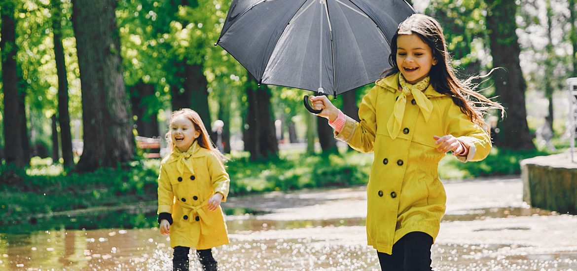 Deux jeunes filles en imperméables jaunes courent joyeusement sous la pluie dans un parc, l'une tenant un parapluie gris. Elles éclaboussent les flaques d'eau en s'amusant.