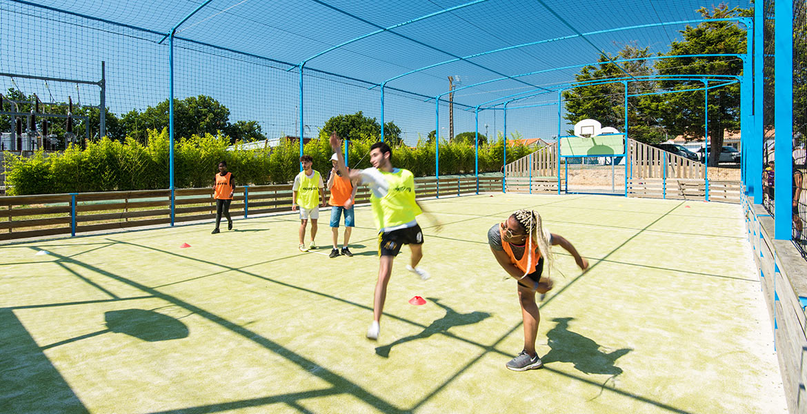 Des enfants jouent au basketball sur un terrain multisport, protégé par un filet bleu sous un ciel ensoleillé.