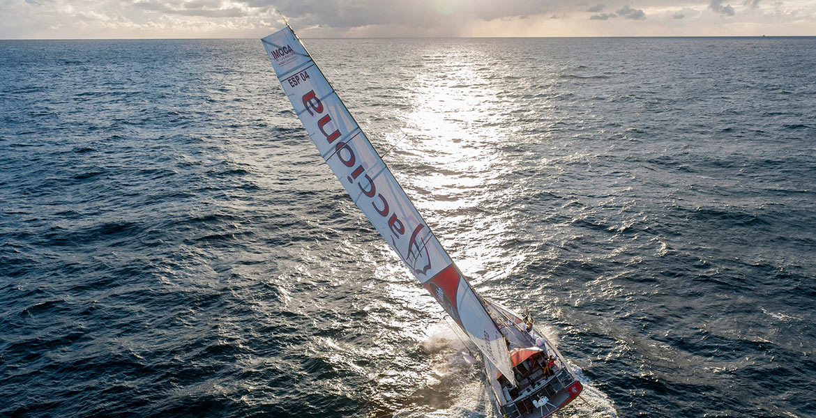 Un voilier de course navigue sur une mer légèrement agitée sous un ciel partiellement nuageux.