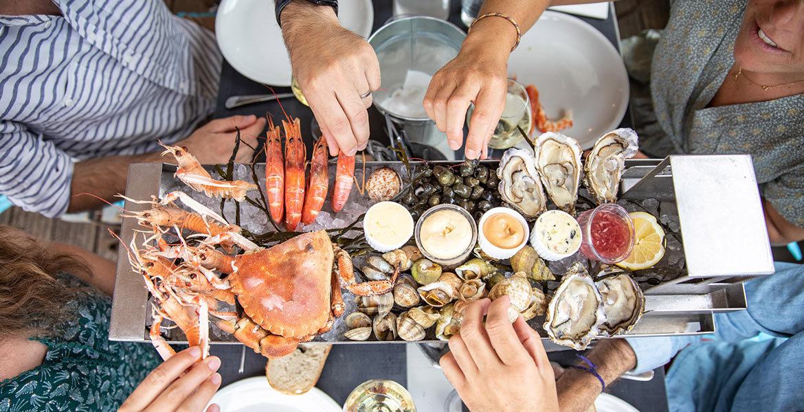 Des personnes autour d'une table dégustant un plateau de fruits de mer comprenant des crevettes, des huîtres, des crabes et divers coquillages, accompagné de sauces et de citron.