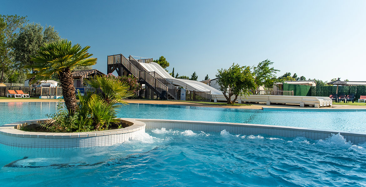 Piscine avec des toboggans blancs en arrière-plan et un îlot de palmiers au centre, entourée de végétation sous un ciel bleu clair.