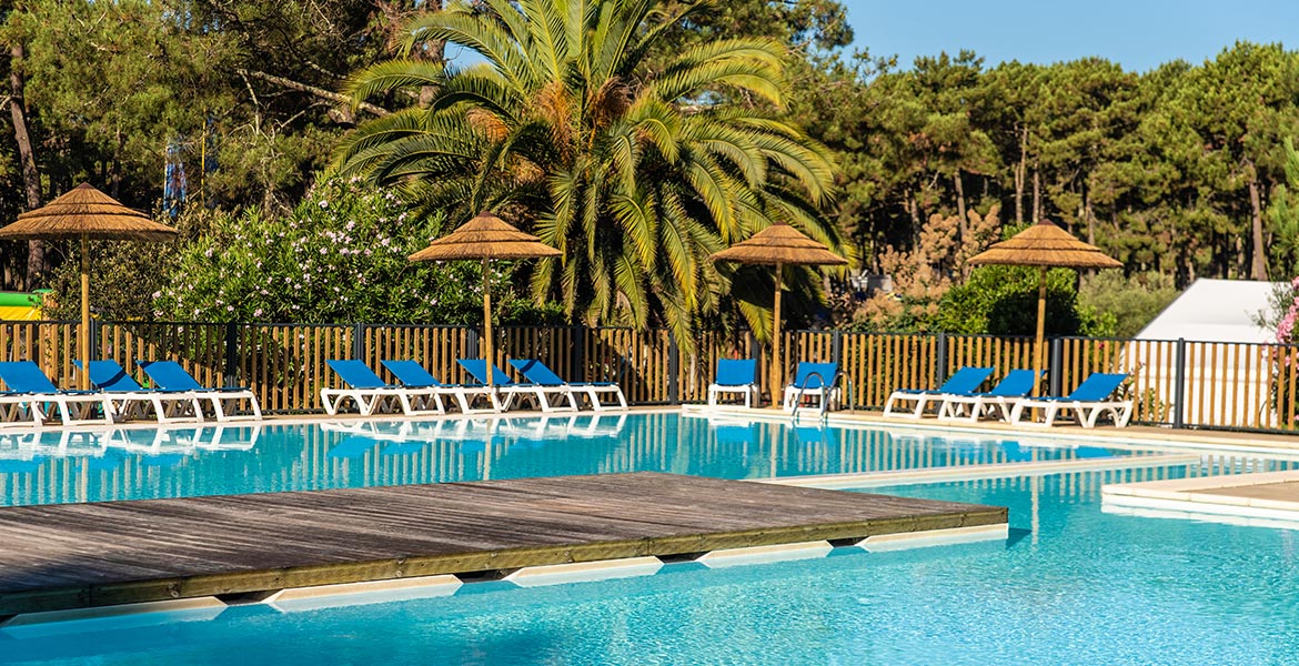 Piscine de style lagon avec une passerelle en bois et des chaises longues bleues sous des parasols en paille, encadrée par une clôture en bois et des arbres.