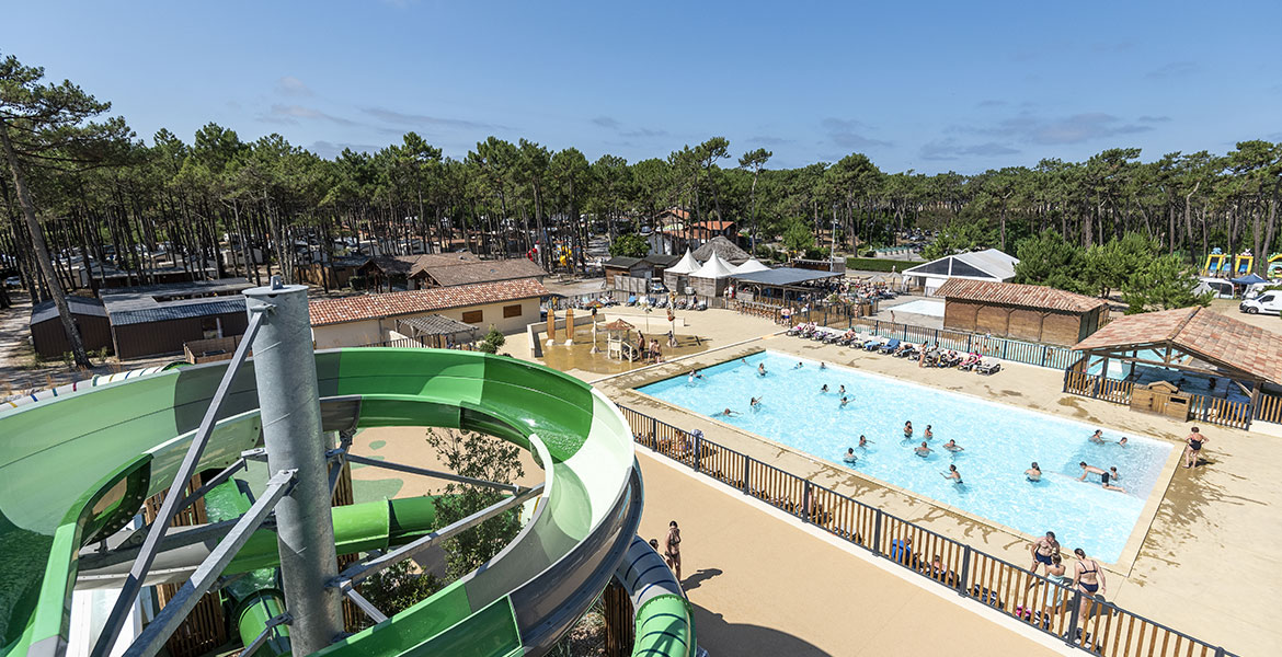 Vue aérienne d'un parc aquatique avec une piscine animée et des toboggans verts, entouré de nombreux arbres et de bâtiments sous un ciel bleu.