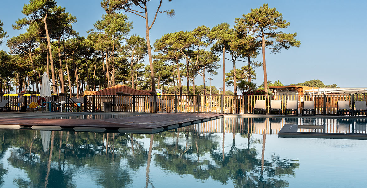 Piscine extérieure calme entourée de chaises longues, avec des arbres en arrière-plan sous un ciel bleu clair.