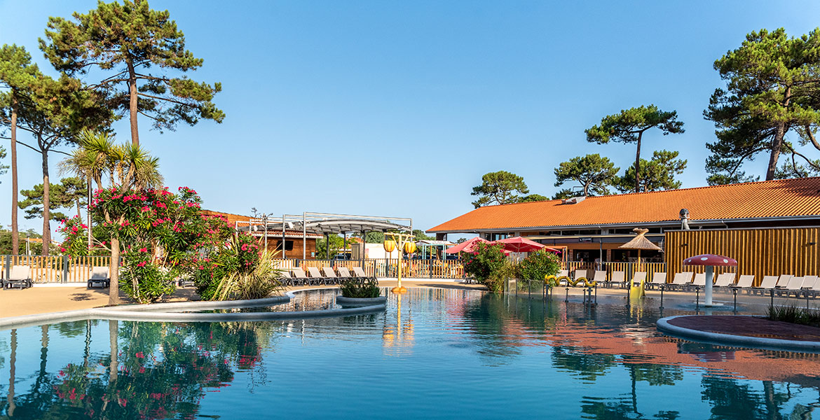 Piscine extérieure dans un complexe de vacances avec chaises longues et parasols sous un ciel bleu, entourée de végétation et de bâtiments à toit rouge.