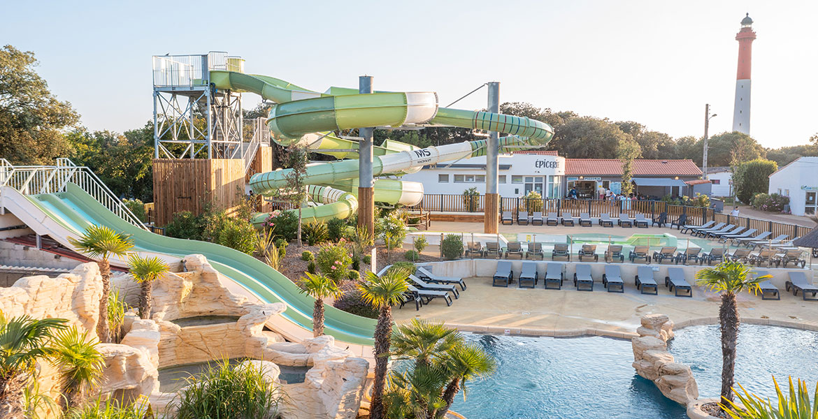 Parc aquatique avec des toboggans verts et une piscine entourée de palmiers, des chaises longues et un phare visible en arrière-plan.