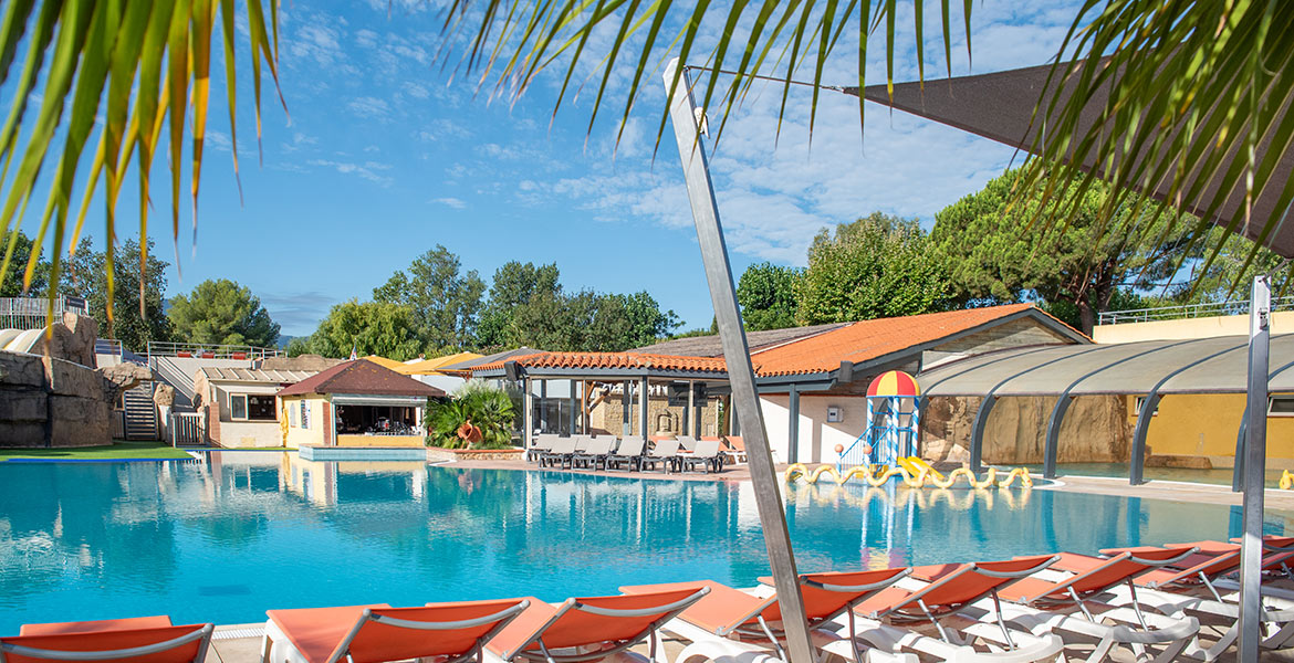 Piscine entourée de chaises longues oranges et de bâtiments à toits rouges, avec des palmiers et des arbres en arrière-plan sous un ciel partiellement nuageux.