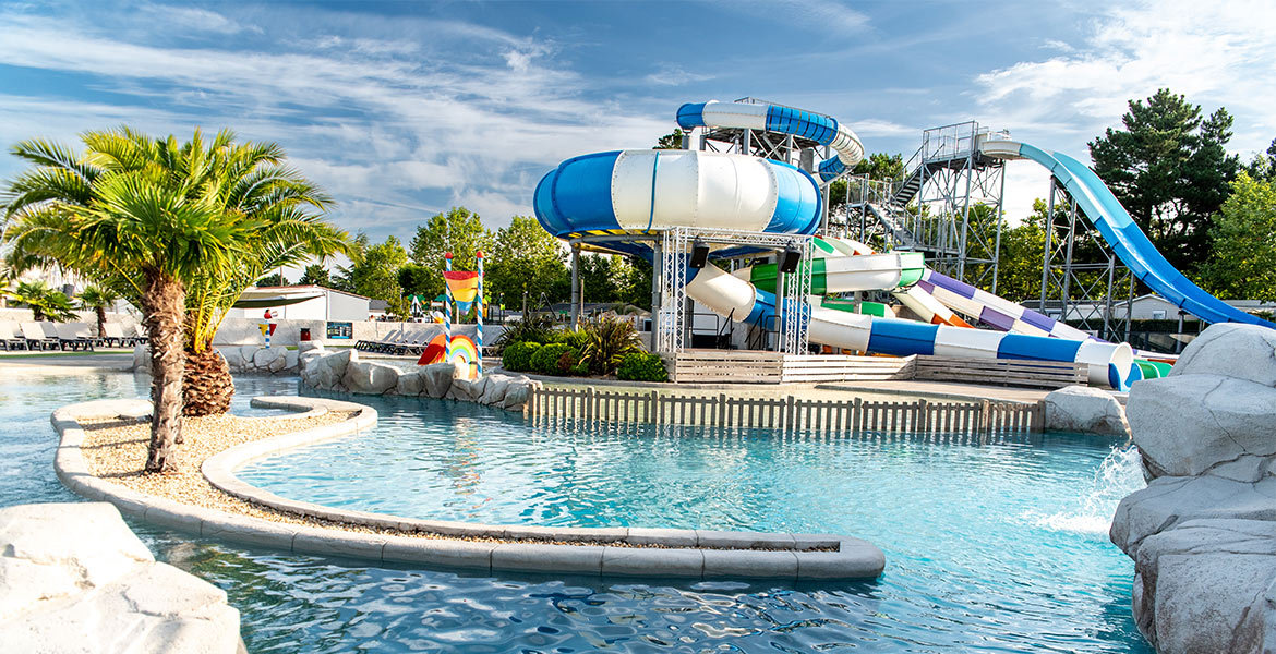 Parc aquatique avec plusieurs toboggans colorés et une grande piscine, entouré de palmiers et de végétation sous un ciel bleu.