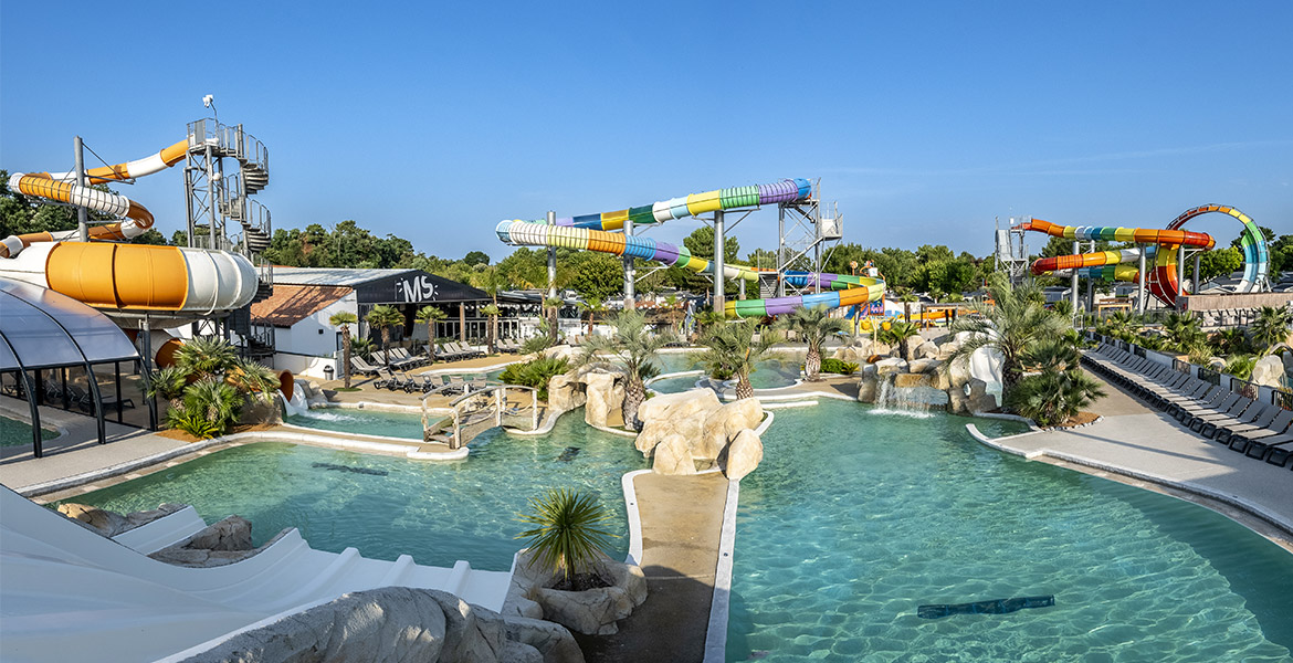 Vue d'ensemble d'un parc aquatique avec de nombreux toboggans colorés, une grande piscine entourée de palmiers et des chaises longues disposées tout autour.