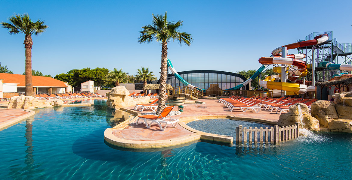 Piscine avec des chaises longues oranges, des toboggans colorés en arrière-plan et des palmiers dispersés autour, sous un ciel bleu clair.