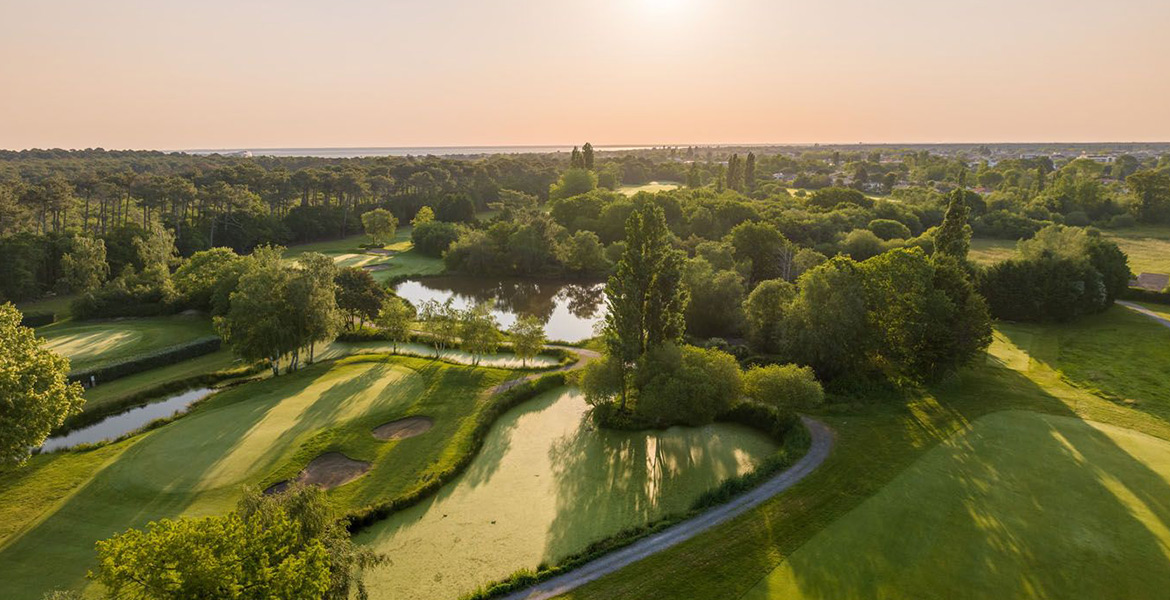 Vue aérienne du golf d'Arcachon au coucher du soleil, montrant des étangs, des bunkers et des allées verdoyantes entourées de forêts.