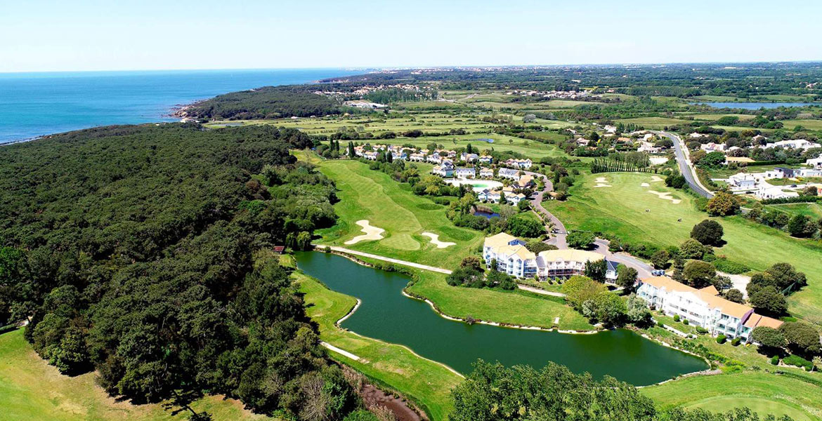 Vue aérienne du Golf de Bourgenay, montrant des fairways verdoyants, des étangs et des bunkers, avec la mer en arrière-plan.