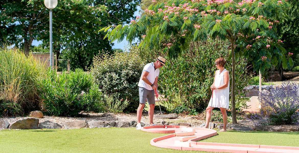 Un couple joue au mini-golf dans un cadre verdoyant au Camping Club MS Le Trianon. L'homme est en train de putter tandis que la femme l'observe, entourés de plantes et d'arbustes colorés.