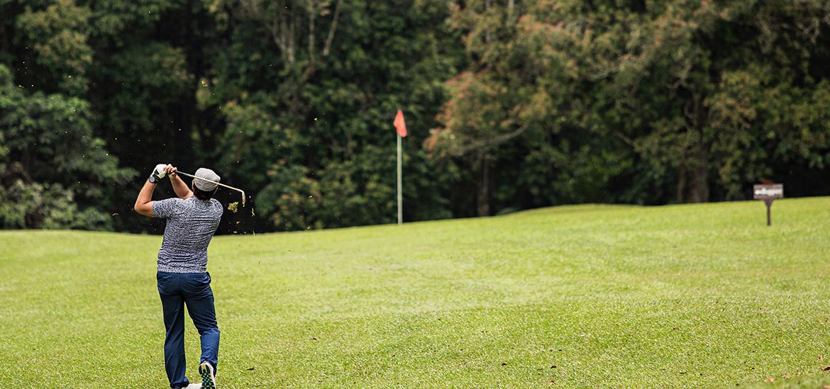 Un golfeur exécute un swing sur un terrain de golf verdoyant avec une forêt en arrière-plan. Un drapeau rouge marque le trou au loin.