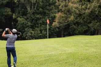 Un golfeur exécute un swing sur un terrain de golf verdoyant avec une forêt en arrière-plan. Un drapeau rouge marque le trou au loin.