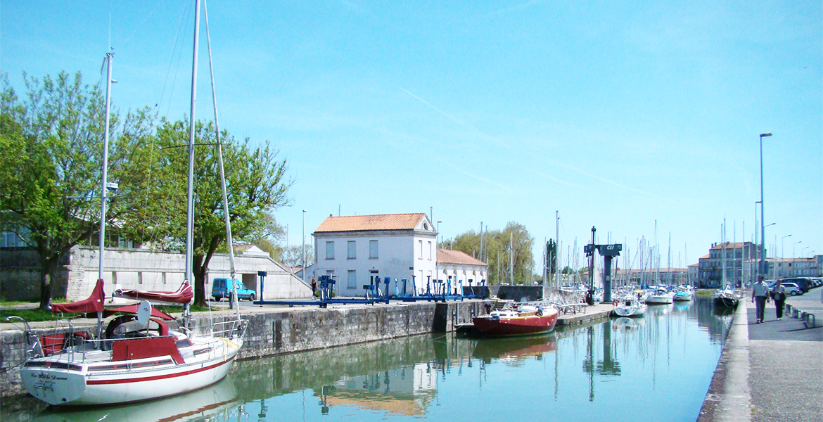 Port de plaisance de Rochefort avec des voiliers amarrés le long d'un canal, bordé par des bâtiments et des arbres sous un ciel bleu.