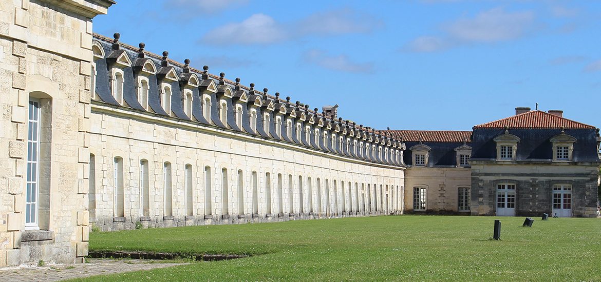 La Corderie Royale de Rochefort. Bâtiment historique en pierre avec de nombreuses fenêtres et un toit mansardé, s'étendant sur un grand terrain vert sous un ciel bleu.