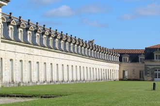 La Corderie Royale de Rochefort. Bâtiment historique en pierre avec de nombreuses fenêtres et un toit mansardé, s'étendant sur un grand terrain vert sous un ciel bleu.