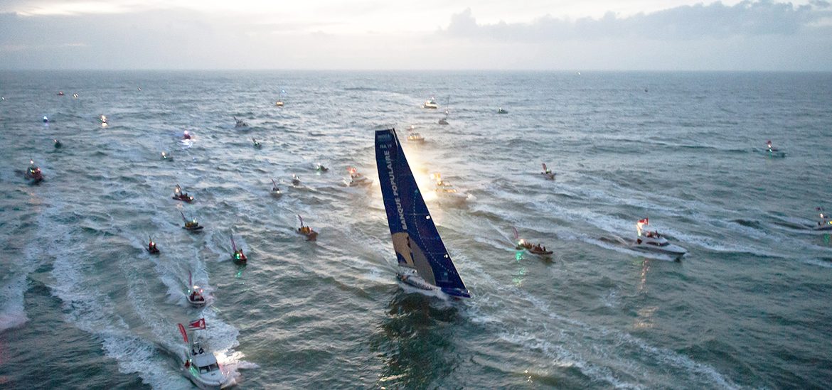 Flotte de bateaux en mer accompagnant un voilier du Vendée Globe au crépuscule.