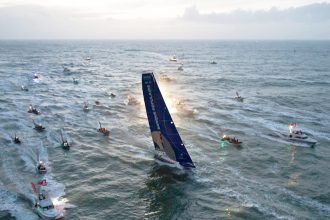 Flotte de bateaux en mer accompagnant un voilier du Vendée Globe au crépuscule.