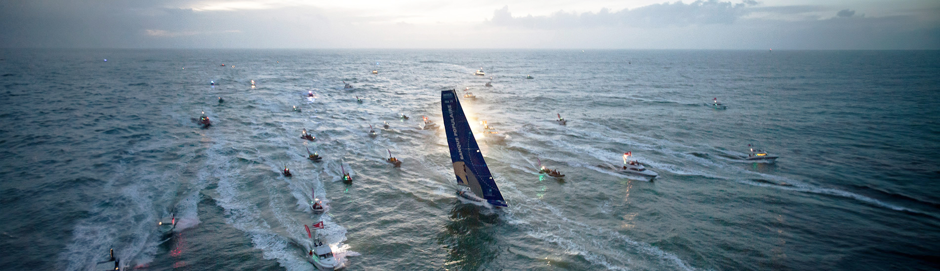 Flotte de bateaux en mer accompagnant un voilier du Vendée Globe au crépuscule.