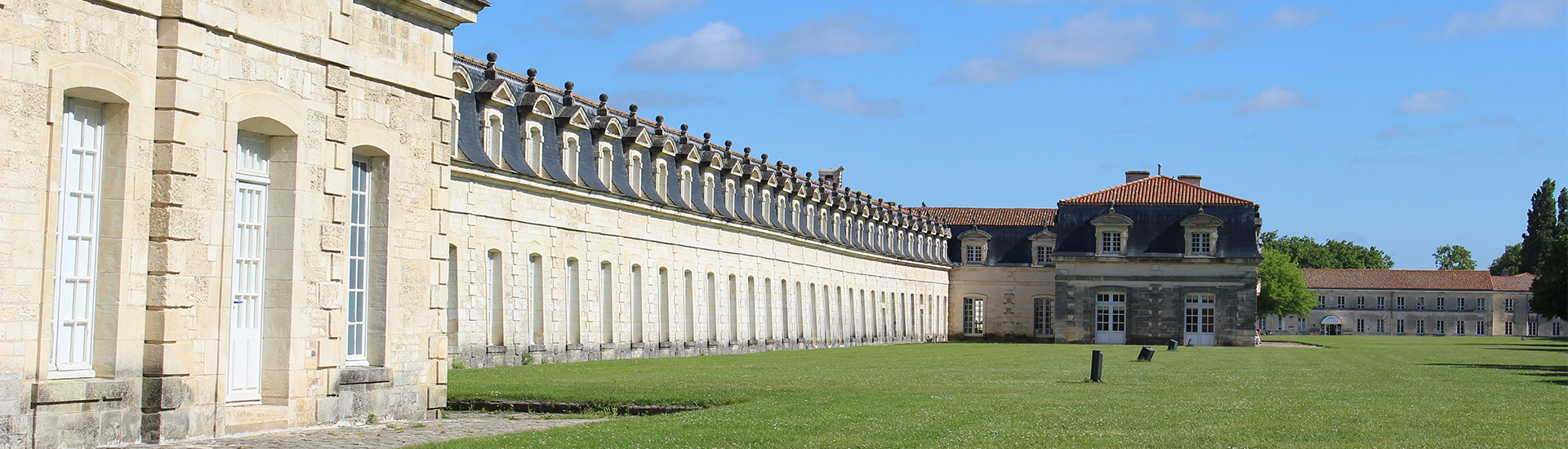 La Corderie Royale de Rochefort. Bâtiment historique en pierre avec de nombreuses fenêtres et un toit mansardé, s'étendant sur un grand terrain vert sous un ciel bleu.