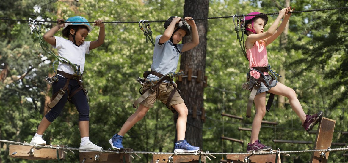 Trois enfants équipés de casques et harnais traversent un parcours d'accrobranche à La Palmyre, en pleine forêt. Ils marchent sur une passerelle suspendue.