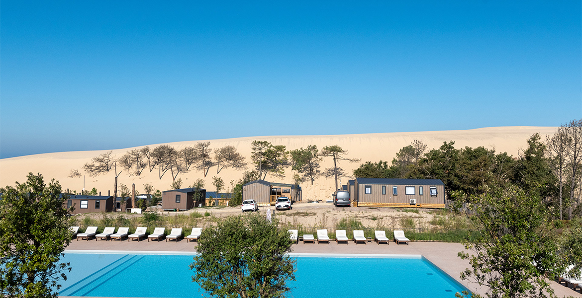 Piscine extérieure du Camping MS Le Pyla 4 étoiles, entourée de végétation et de transats, avec la Dune du Pilat en arrière-plan sous un ciel bleu clair.