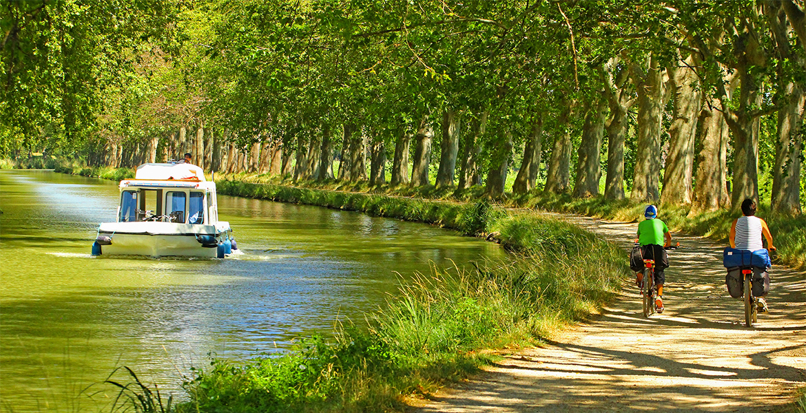 Deux cyclistes roulent sur un chemin ombragé longeant le canal du midi bordé d'arbres, tandis qu'un bateau navigue doucement sur l'eau.