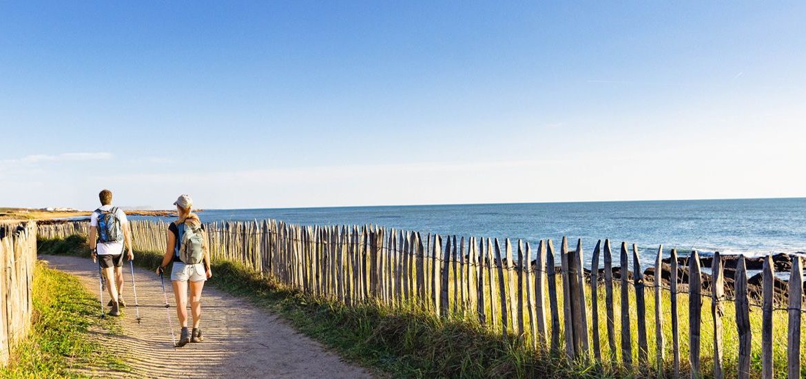 Deux randonneurs marchent sur un sentier côtier bordé d'une barrière en bois, avec vue sur la mer sous un ciel dégagé.