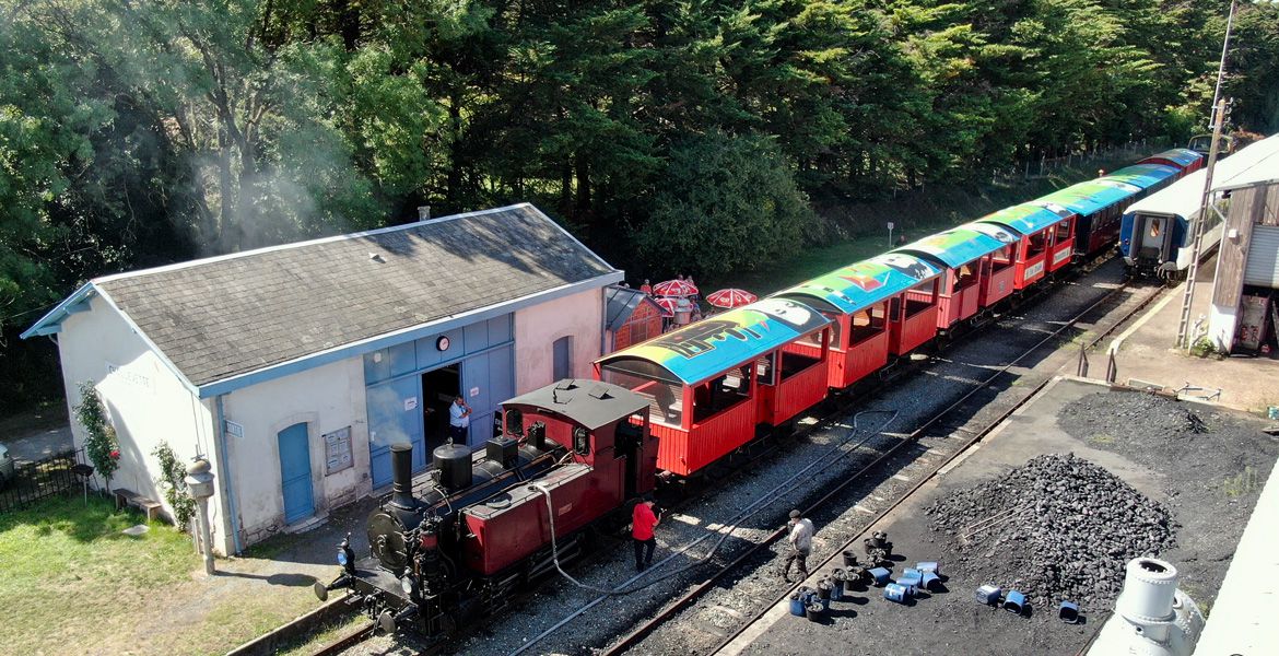 Vue aérienne d’un train touristique rouge à vapeur, le Train des Mouettes, stationné près d’une petite gare entourée de végétation. Des seaux de charbon sont disposés au sol à proximité de la locomotive.