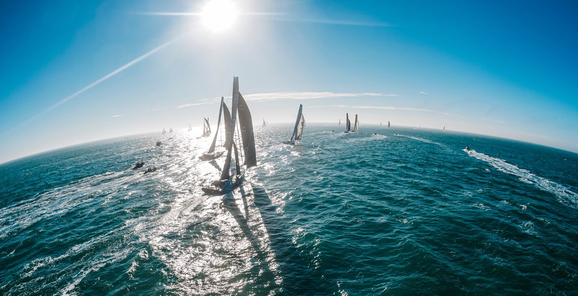 Des voiliers du Vendée Globe, aux Sables d'Olonne, naviguent sur une mer scintillante sous un ciel bleu éclatant. Le soleil brille intensément, projetant des reflets lumineux sur l'eau.