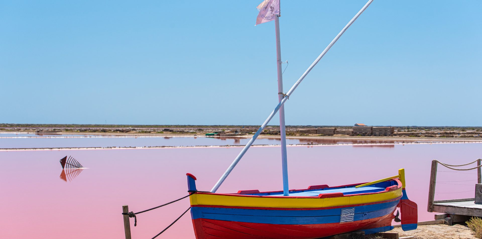 Un bateau de pêche coloré est amarré sur la rive d'un marais salant aux eaux roses sous un ciel bleu.